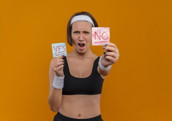 字身穿运动服 头箍的年轻健身女士站在橙色的墙上 兴高采烈地展示着两张写着“是”和“不是”的提醒纸年轻人纸人