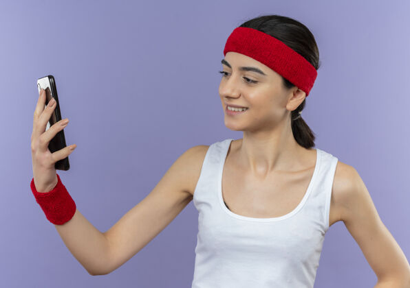 头带身穿运动服 头戴头巾的年轻健身女士站在紫色的墙上 看着智能手机的屏幕 自信地微笑着微笑运动年轻