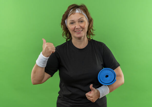 运动身穿黑色t恤 头箍 手持瑜伽垫的中年运动女性站在绿色的墙壁上愉快地微笑着竖起大拇指垫子瑜伽抱着