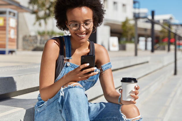 眼镜快乐潮人的户外镜头在网站上看评论 聚焦在手机屏幕上 端着外卖咖啡 坐在楼梯上 穿着牛仔服 在漫游中享受高速互联网工作服短信短信