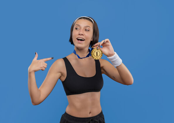 手指身穿运动服的年轻女子 脖子上挂着金牌 用食指指着奖牌微笑自信地站在蓝色的墙上颈部人物自信