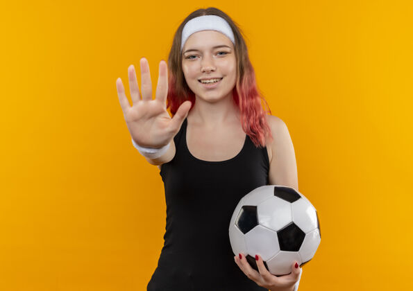 人身着运动服的年轻健身女士手持足球 站在橙色的墙上微笑着示意停车抱微笑年轻