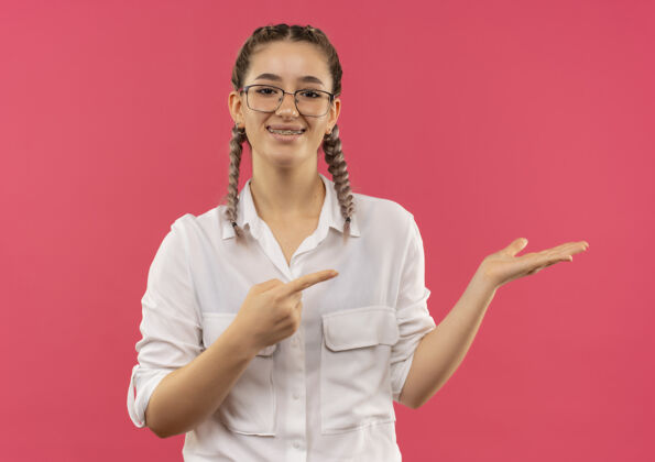 指着戴着眼镜 梳着辫子 穿着白衬衫的年轻女学生面带微笑地站在粉红色的墙上 用手指指着旁边的东西手臂市民衬衫