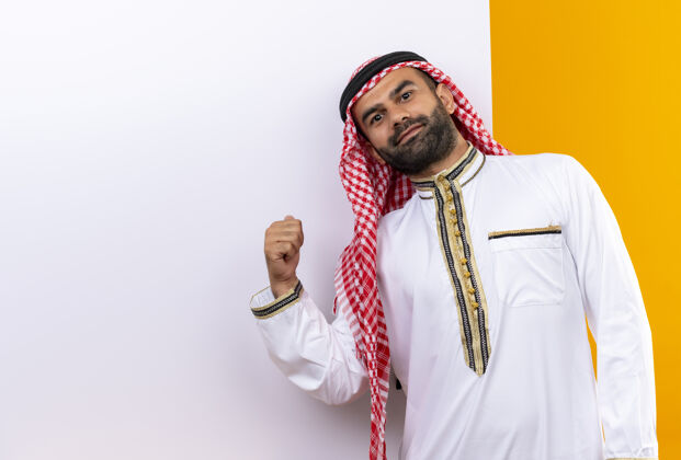 手指身着传统服装的阿拉伯商人站在空白的广告牌旁 手指指着广告牌 脸上带着自信的微笑 越过橙色的墙壁传统脸微笑