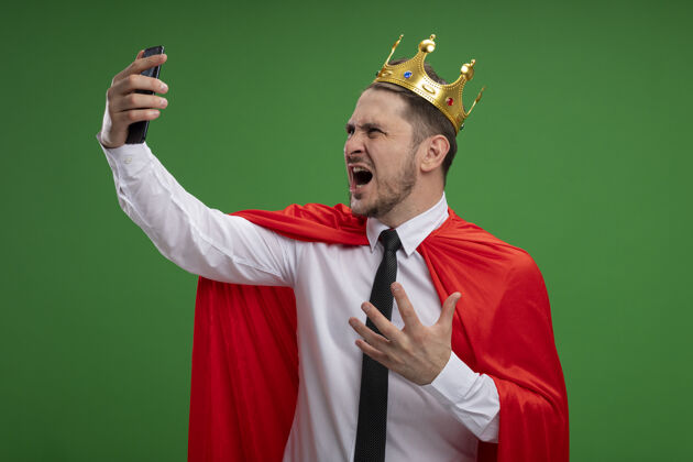 商务人士穿红斗篷戴皇冠自拍的超级英雄商人用智能手机疯狂愤怒地站在绿色背景下斗篷愤怒超级