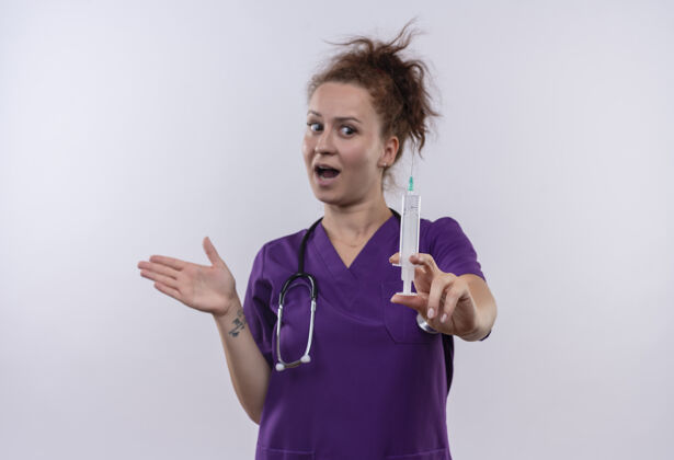 女人年轻的女医生穿着医疗制服 手持听诊器和注射器 站在白色的墙上 看上去很惊讶拿着惊讶听诊器