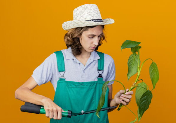 持有年轻的园丁 穿着连体衣 戴着帽子 用树篱剪剪着植物帽子削减男人