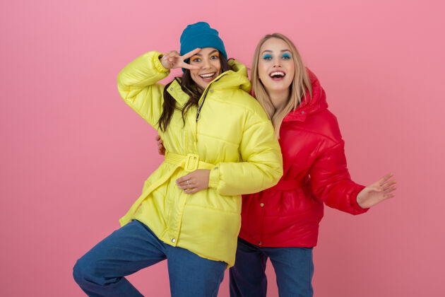肖像两位迷人的活跃女性穿着鲜艳的红黄相间的冬季羽绒服在粉色的墙上摆着姿势 朋友们在一起玩得很开心 温暖的外套时尚潮流 疯狂搞笑的面孔服装穿着女人