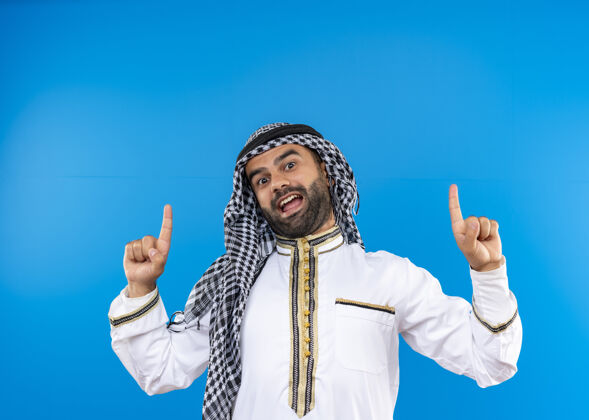 传统一位身着传统服装的阿拉伯男子站在蓝色的墙上 手指朝上 微笑着男人穿手指
