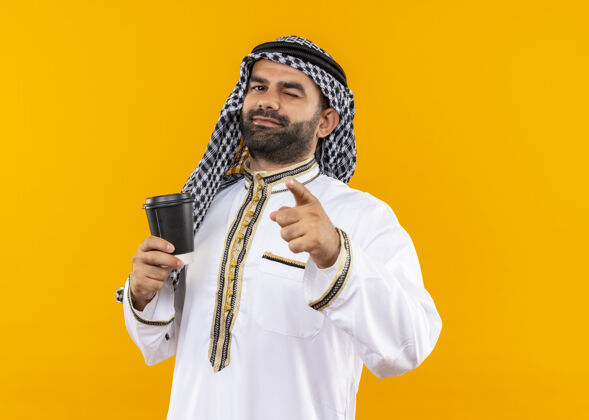 咖啡身着传统服装的阿拉伯商人手持咖啡杯 手指眨着眼睛 微笑着站在橙色的墙上穿手指眨眼