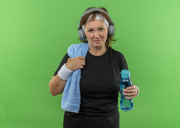 女人中年运动女性 身穿黑色t恤 头上系着头带 脖子上系着毛巾 手里拿着一瓶水 劳累而快乐地站在绿色的墙上毛巾中瓶子