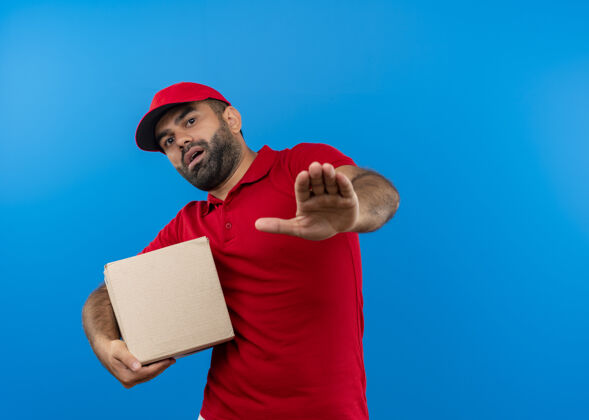 使留着胡须的送货员穿着红色制服 戴着帽子 拿着盒子 站在蓝色的墙上 一副恐惧的表情恐惧交货停止