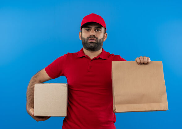 拿着一个留着胡子的送货员 穿着红色制服 戴着帽子 拿着盒子和纸包装 严肃的脸站在蓝色的墙上制服交货帽子
