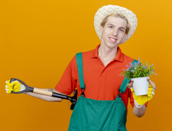男人年轻的园丁穿着连体衣戴着帽子拿着铲子展示盆栽连身衣穿花盆