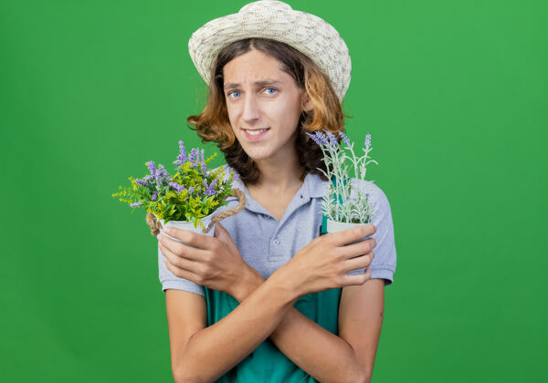 植物年轻的园丁穿着连体衣戴着帽子拿着盆栽微笑着穿着抱着站着