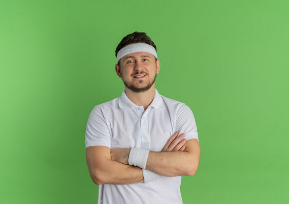 人身穿白衬衫 头箍 双手交叉放在胸前 自信地站在绿色墙壁上的年轻健身男子头带十字架表情
