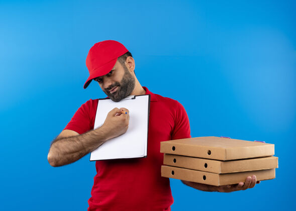 披萨满脸胡须 身穿红色制服 头戴鸭舌帽的送货员站在蓝色的墙上 手里拿着披萨盒和空白页要求签名的剪贴板页面盒子胡子
