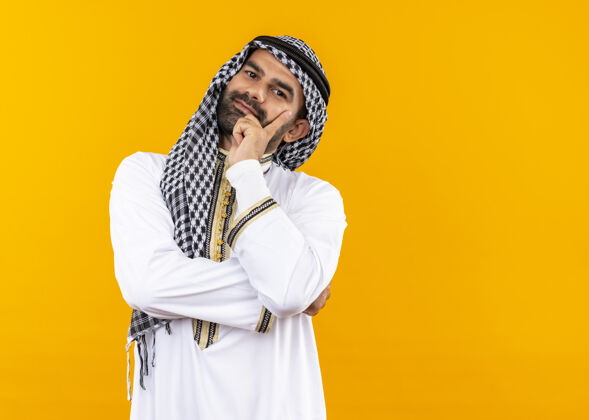 积极身着传统服饰的阿拉伯商人站在橙色的墙上 脸上挂着快乐而积极的微笑阿拉伯语站着商人