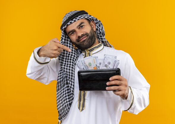 手指身着传统服饰的阿拉伯商人手里拿着钱包 手里拿着现金 微笑着自信地用手指着站在橙色墙上的现金现金阿拉伯语看