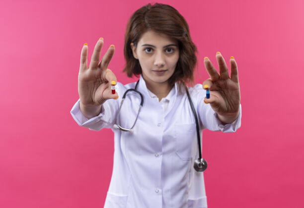 严肃身穿白大褂 手持听诊器的年轻女医生站在粉红色的墙上 表情严肃 手里拿着药片药片听诊器年轻人