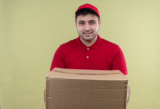 拿着年轻的送货员穿着红色制服 戴着帽子拿着盒子 友好地站在绿色的墙上微笑着站着看着送货
