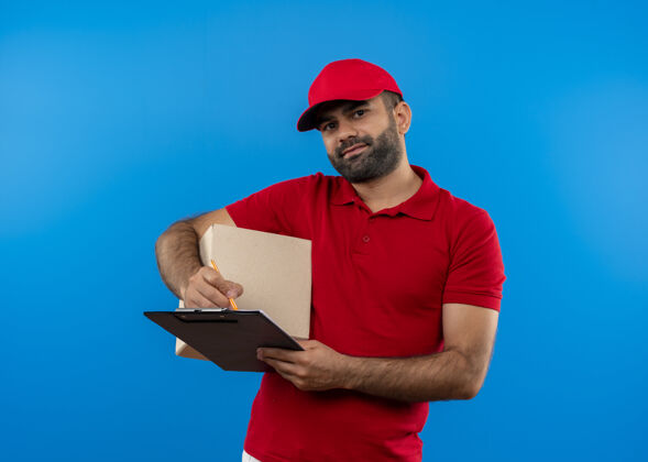 帽子留着胡子的送货员穿着红色制服 戴着帽子 拿着盒子 微笑着自信地站在蓝色的墙上制服看着微笑