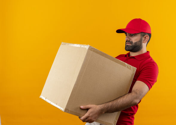 重量留着胡须的送货员穿着红色制服 戴着帽子 手里拿着一个大箱子 站在橘黄色的墙上 承受着沉重的重量拿着送货包装