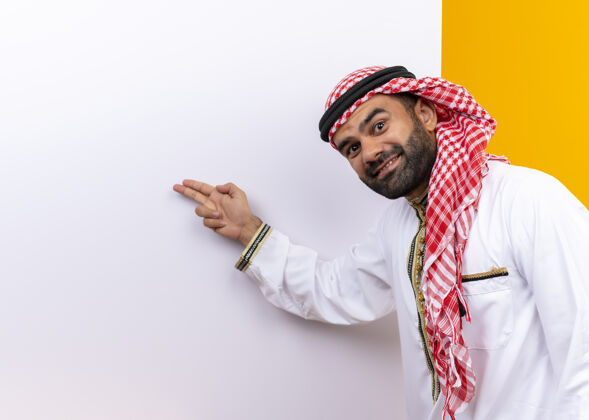 商务人士身着传统服装的阿拉伯商人站在空白的广告牌旁 用手指着它 脸上带着微笑越过橘色的墙壁脸传统阿拉伯语