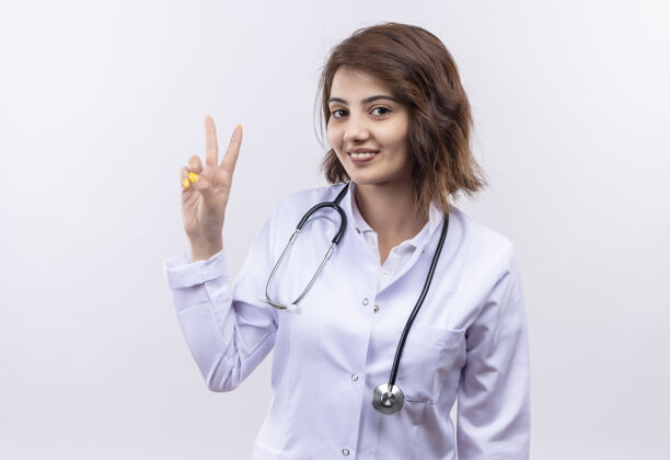 站着身穿白大褂 手持听诊器的年轻女医生站在白墙上 手指朝上 二号微笑着站着手指号码年轻人
