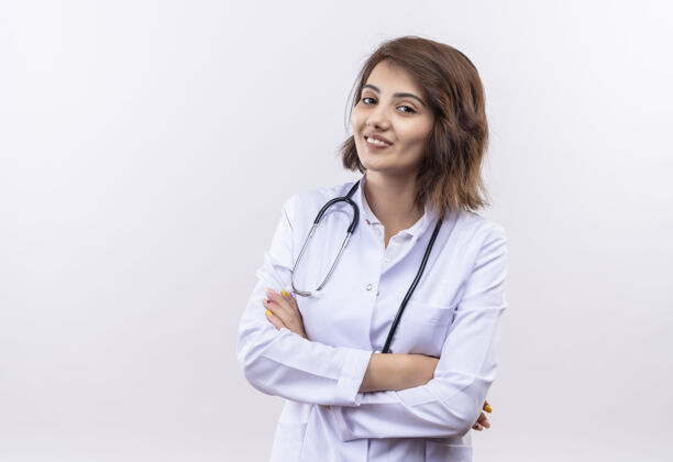 女人身穿白大褂 手持听诊器的年轻女医生微笑着自信地站着 双臂交叉在白墙上微笑医生胳膊