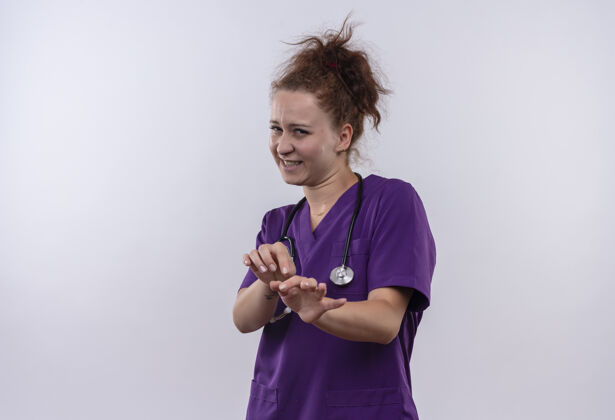 制服年轻的女医生穿着医疗制服 手持听诊器 站在白墙上 用厌恶的表情做着自卫的手势站立穿着防御