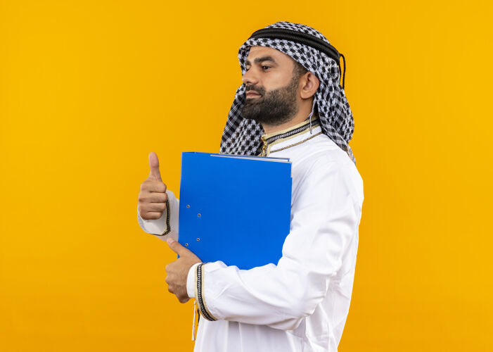 商务人士身着传统服装的阿拉伯商人手拿蓝色文件夹 竖起大拇指 站在橙色的墙上阿拉伯语传统拇指
