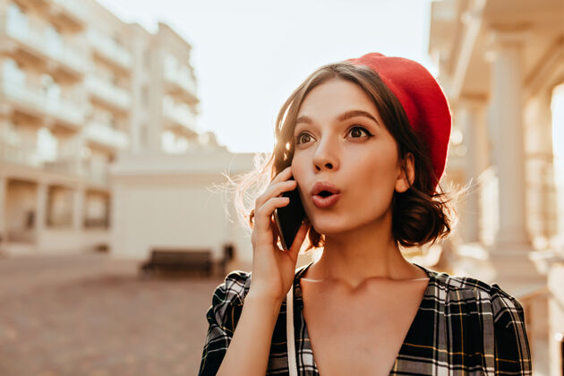 优雅一个戴着红帽子的惊喜女孩在打电话一个漂亮的黑发女士拿着智能手机在阳光明媚的秋天摆姿势卷发年轻漂亮
