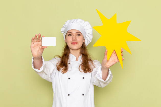 穿女厨师身穿白色厨师服 手持黄色标志和绿色墙壁上的卡片的正面视图前面女厨师护士