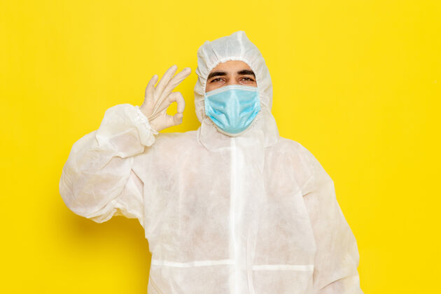 外套身穿白色特殊防护服 带面罩的男性科学工作者正面图 黄色墙上显示“好”标志危险科学科学