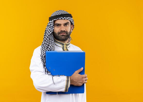 商务人士身着传统服饰的阿拉伯商人手持蓝色文件夹 自信地微笑着站在橙色的墙上阿拉伯语文件夹穿