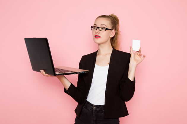 前面穿着黑色夹克的女上班族在粉色墙上用手提电脑持卡的前视图办公室持有工作