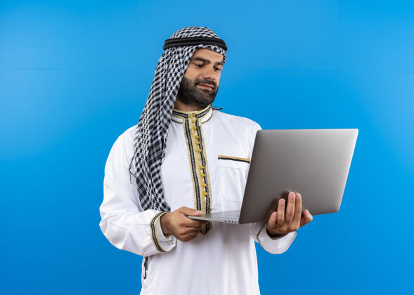 微笑身着传统服装的阿拉伯商人拿着笔记本电脑站在蓝色的墙上面带微笑地看着它阿拉伯语笔记本电脑传统