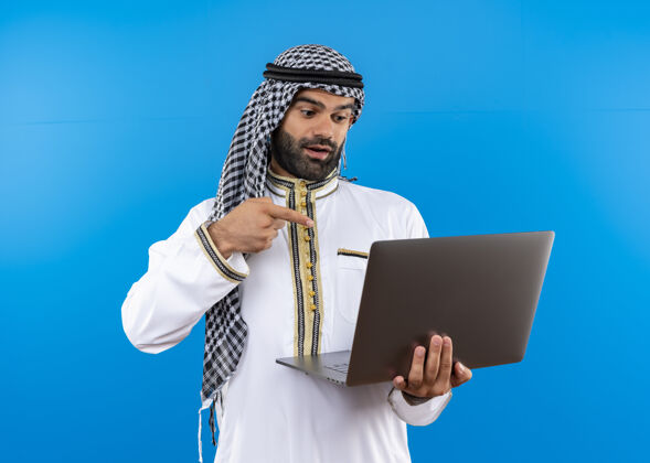 阿拉伯语身着传统服装的阿拉伯商人手持笔记本电脑 面带微笑地看着它 手指指着它站在蓝色的墙上穿手指商务人士