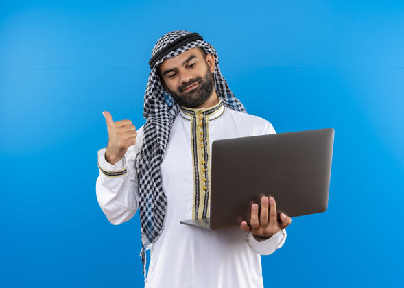 传统身着传统服装的阿拉伯商人拿着笔记本电脑看着他的笔记本电脑屏幕 脸上带着微笑站在蓝色的墙上看背屏幕