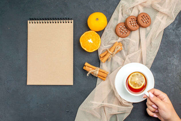 棍棒俯视图一杯茶 柠檬在女性手中肉桂棒饼干在米色披肩橙色记事本在黑暗的表面饼干景观肉桂