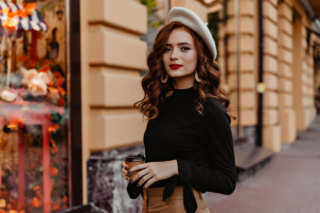 户外穿着棕色贝雷帽的美丽的欧洲女孩在户外消磨时光好奇的法国姜女士站在街上喝茶卡布奇诺街头帽子