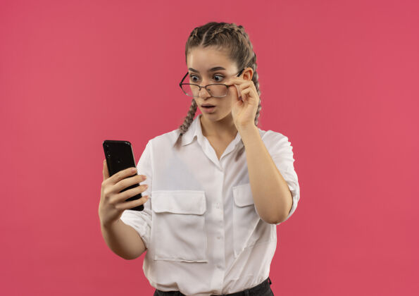 人戴着眼镜 梳着辫子 穿着白衬衫的年轻女学生站在粉红色的墙上 看着手机屏幕 既惊讶又惊讶站姿势年轻人