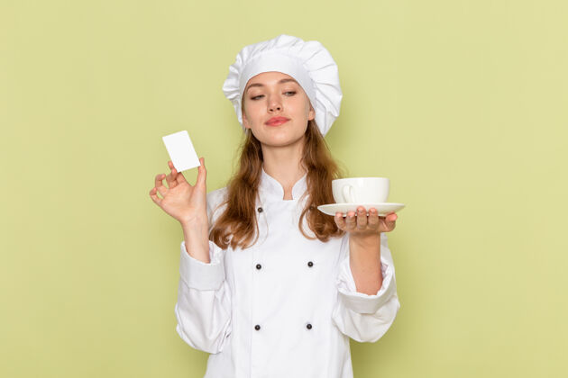 杯子身着白色厨师服的女厨师手持白色塑料卡和杯子站在绿色墙上的正视图烹饪套装女厨师