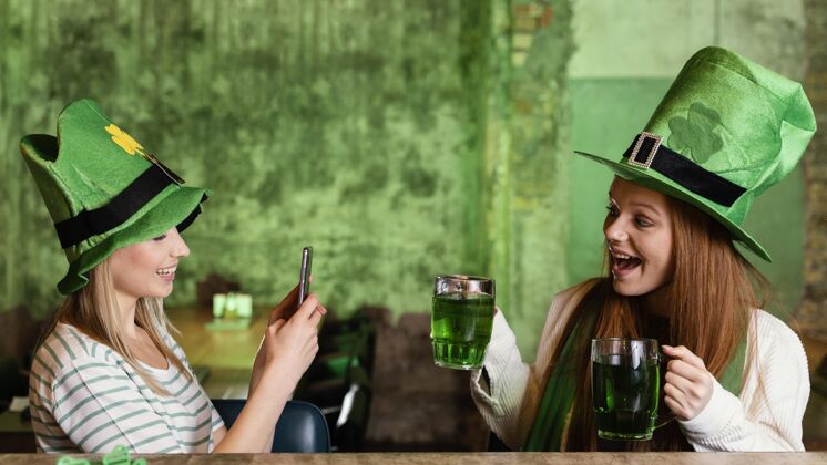 酒吧快乐的女性朋友一起庆祝圣帕特里克节智能手机和饮料手机三叶草圣帕特里克日