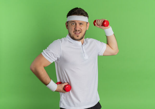 手势身穿白衬衫 头箍戴着哑铃的年轻健身男子站在绿色的墙壁上紧张而自信地锻炼身体站立工作姿势