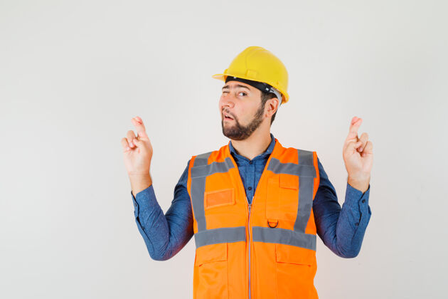 车间年轻的建筑工人穿着衬衫 背心 头盔保持手指交叉和眨眼 前视图帽子工业工匠