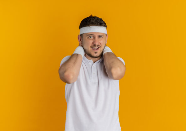小伙子身穿白衬衫 头戴头巾的年轻健身男子站在橙色的墙上 看着前方感到不适运动人看