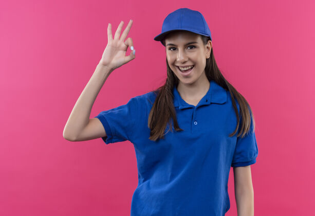 Ok身穿蓝色制服 头戴帽子的年轻送货女孩看着卡姆拉 开心地微笑着 露出“ok”的手势站立表情标志