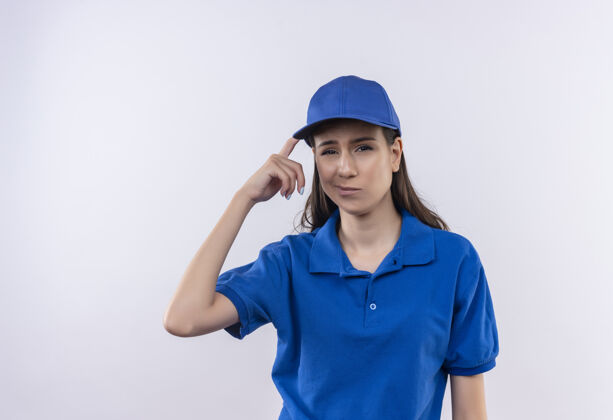 年轻身穿蓝色制服 戴着帽子的年轻送货女孩看着摄像机困惑地指着自己的太阳穴找错了 忘了太阳穴看指向